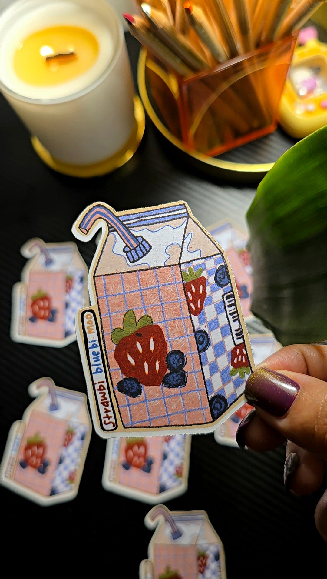 Hand holding a decorative milk carton card with strawberries and blueberries, surrounded by candles and matches.