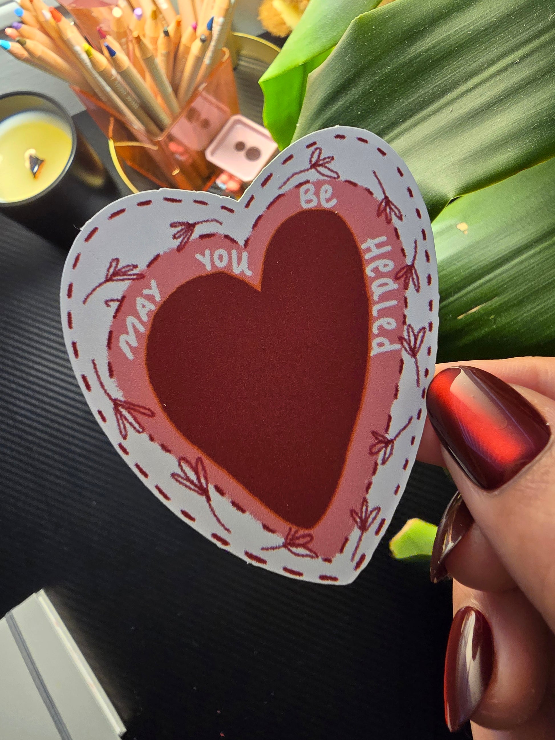 Heart-shaped card with text held by a hand with red nail polish, against a blurred background.