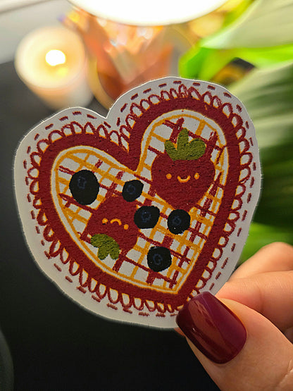 "Heart-shaped waffle die-cut sticker with kawaii strawberry and blueberries, held by a hand with dark red nail polish."