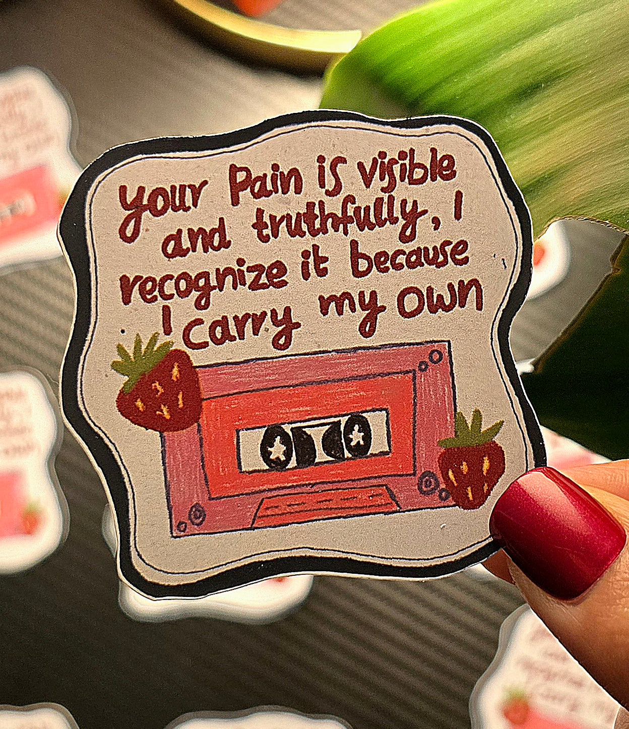 Sticker with a cassette tape design and text held by a hand with red nail polish.