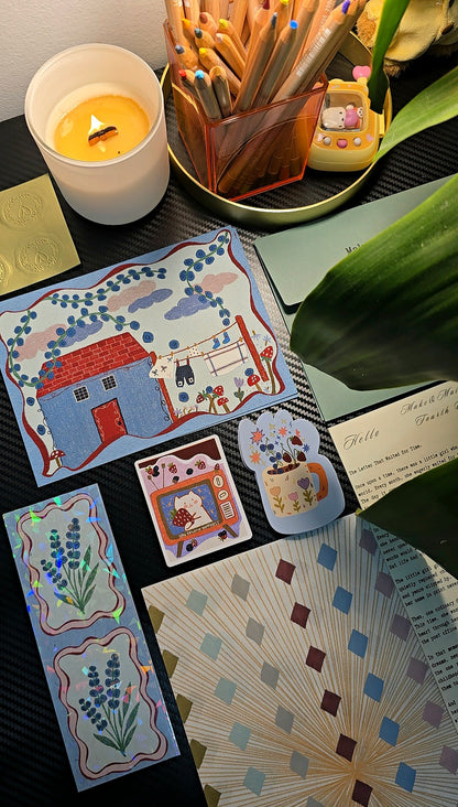 A styled flat-lay of handmade illustrated cards and bookmarks on a dark textured surface, surrounded by a lit candle, a pencil holder, and a green plant leaf.