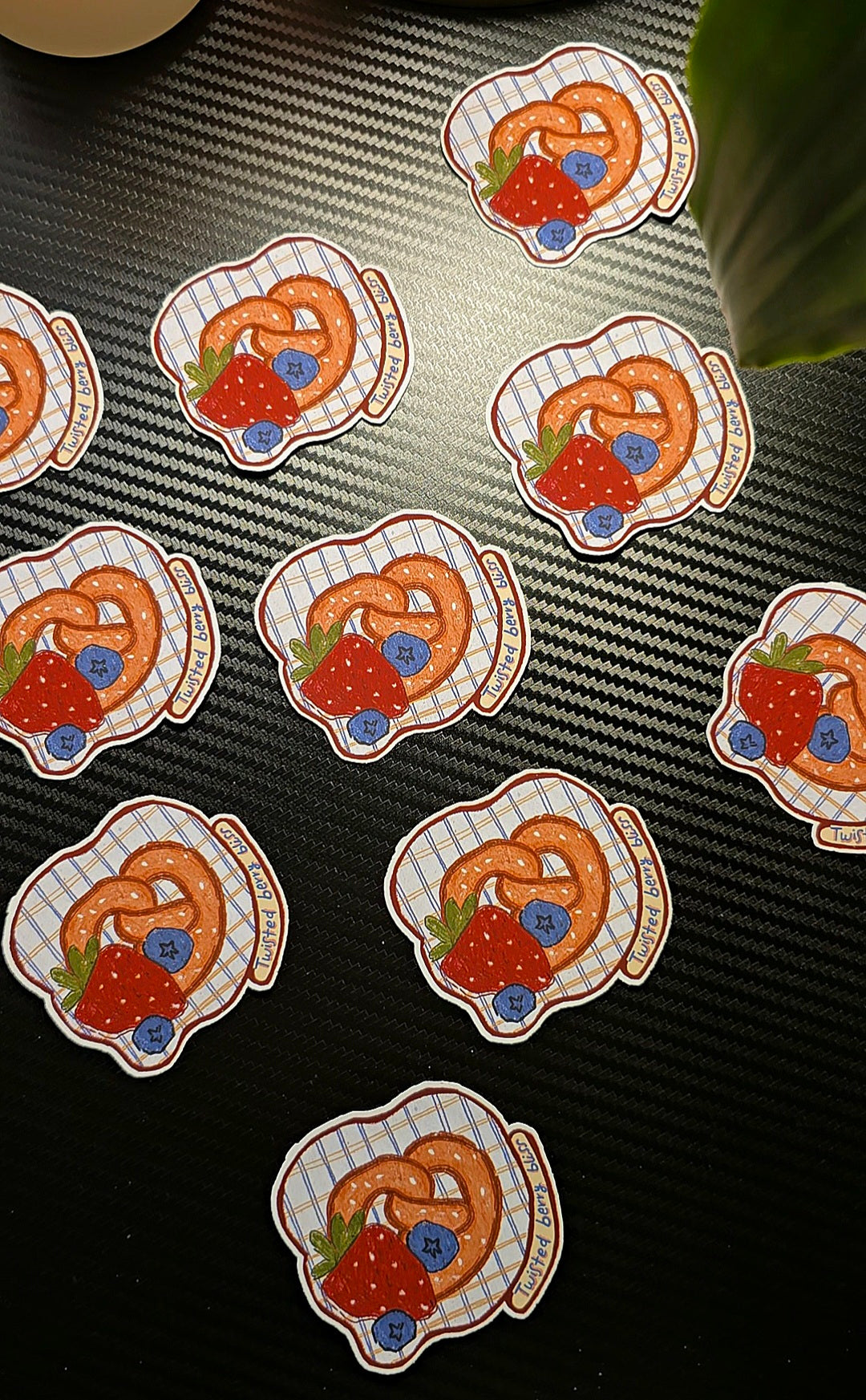 Multiple identical round stickers featuring a pretzel with strawberries and blueberries on a white checkered plate, arranged on a dark textured surface.