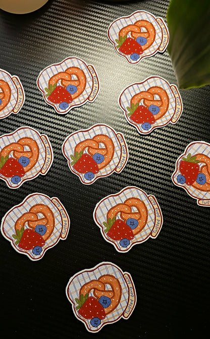 Multiple identical round stickers featuring a pretzel with strawberries and blueberries on a white checkered plate, arranged on a dark textured surface.